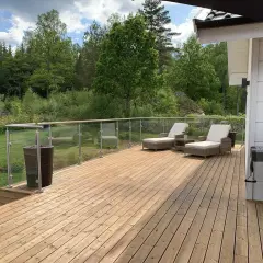 Träaltan med solstolar och grill, omgiven av grönskande natur. Ett nymålat staket syns i bakgrunden.
