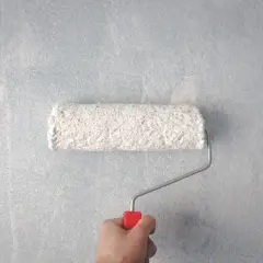 En hand håller en färgroller med vit färg och målar på en ljusblå, texturerad tapet.