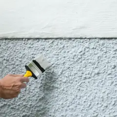 Hand målar en vägg utomhus med en pensel. Det är viktigt att tänka på luftfuktighet när man ska måla utomhus för bästa resultat.