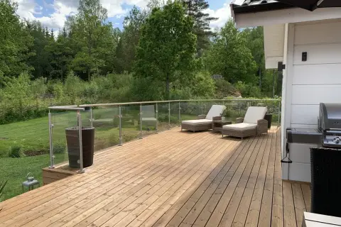 Träaltan med solstolar och grill, omgiven av grönskande natur. Ett nymålat staket syns i bakgrunden.