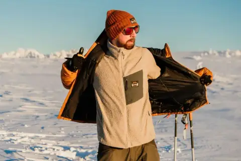 Man i orange mössa och solglasögon visar upp sin lager på lager-klädsel i snölandskap med skidstavar.